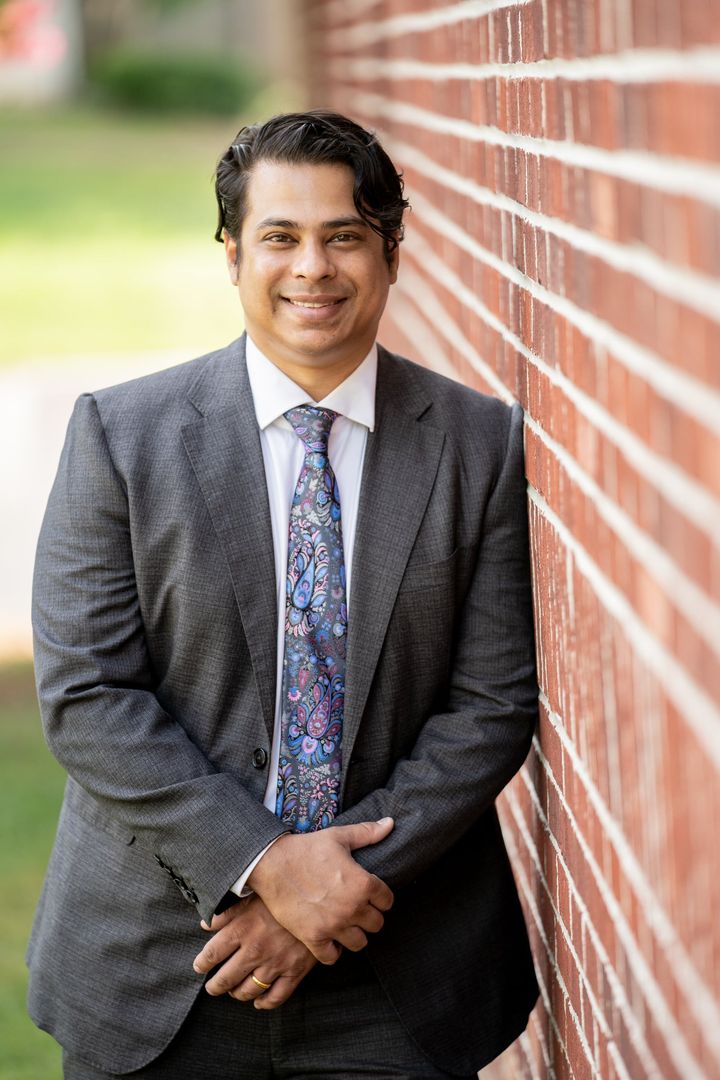 Shree Mulay in a charcoal suit and patterned tie, smiling and leaning against a red brick wall outdoors.